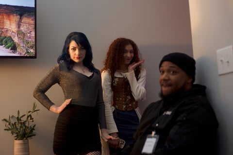 Three women posing together indoors. Woman on left wears brown and black. Woman in middle wears patterned vest over white shirt. Woman on right wears black jacket. A plant and framed landscape photo visible in background.