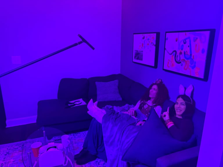 Three people sitting on a black couch in a room lit with purple neon lights, gesturing while watching screens on the wall
