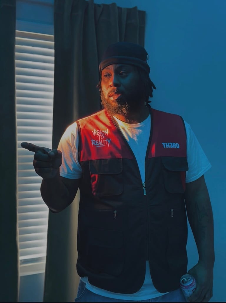 Man in black and red tactical vest with cap pointing at camera against blue lighting and window blinds