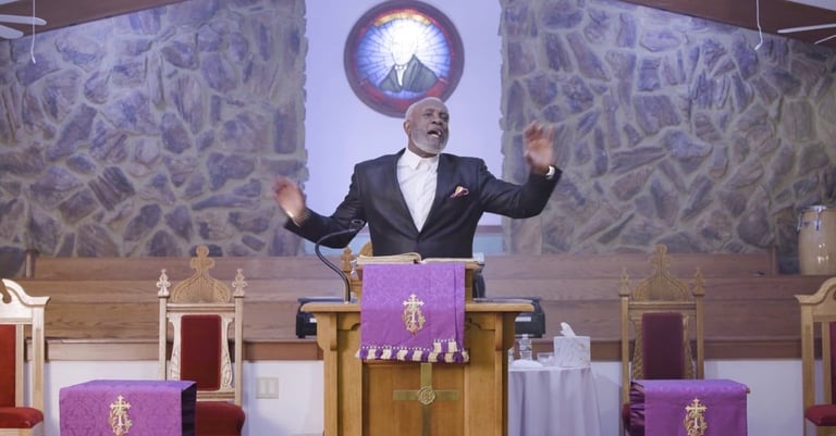 Priest in formal attire speaking from a church pulpit with arms raised, purple religious cloth draped on furniture, stained glass window above