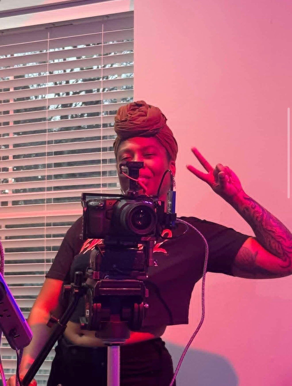 Videographer holding professional camera in front of pink wall with window blinds in background