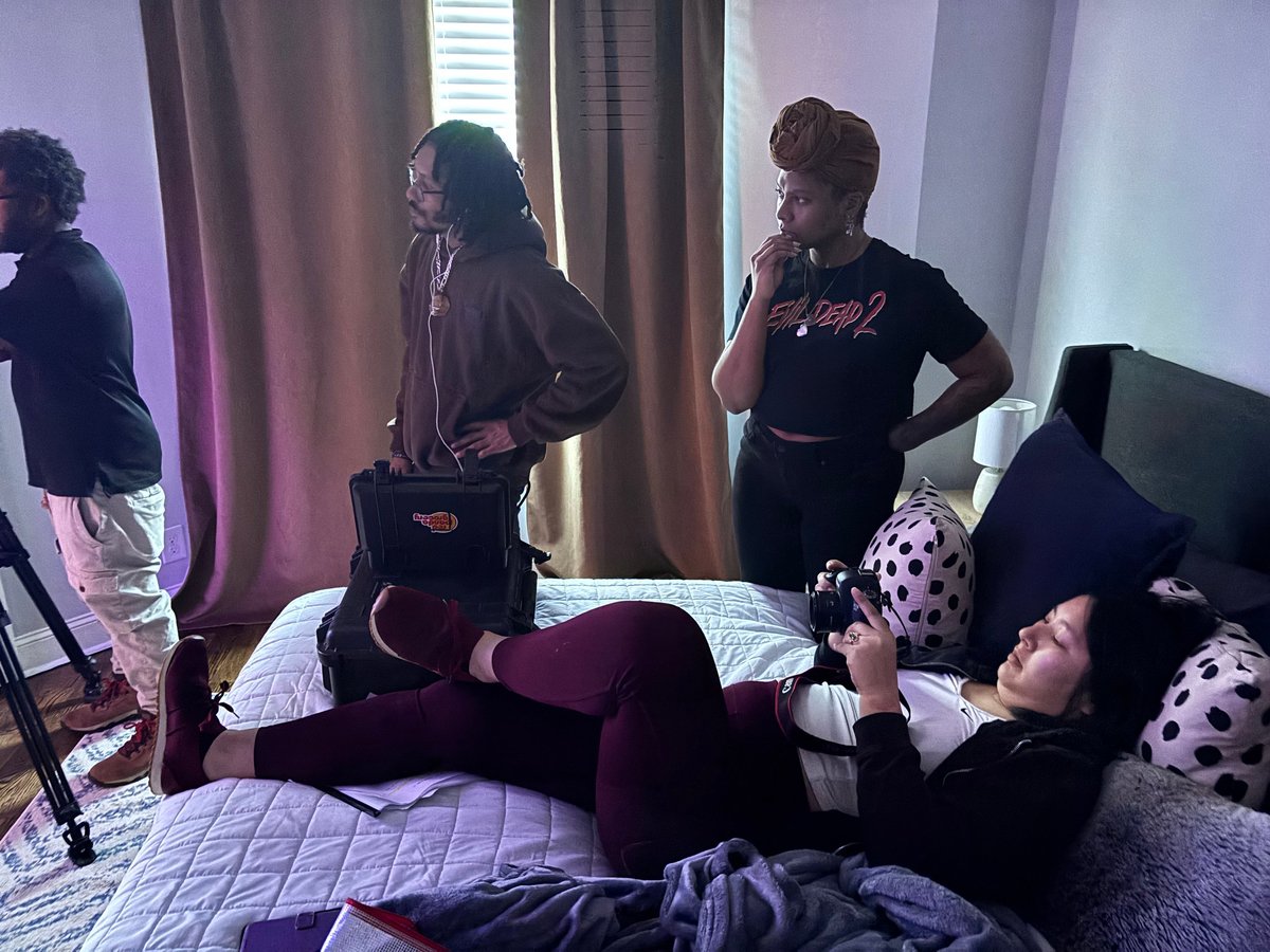 Group of people in a bedroom with one person lying on a bed and others standing nearby in casual conversation
