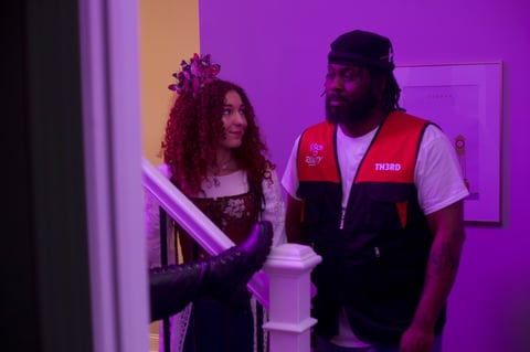 Two people in an indoor setting with purple lighting. Woman on left wearing dark dress with red flower crown, man on right wearing red and black vest over white shirt.
