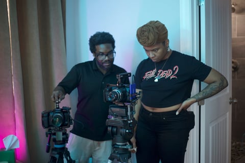 Two people operating professional film cameras in a studio with pink and blue neon lighting