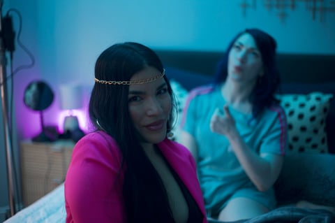 Woman in magenta blazer and chain headpiece sitting on bed with neon blue and pink lighting, another woman visible in background